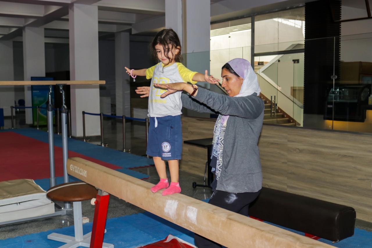 A young girl wearing a yellow t-shirt and blue shorts practices on a balance beam at IVY STEM International School. She is guided by an adult wearing a hijab, who closely observes and assists her. The indoor setting suggests a gymnastics training facility or sports center at IVY STEM International School.